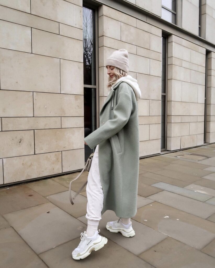 A girl with a sage green trench coat on and some balenciaga triple s sneakers on the girl is Standing in front of of a brick building and on the side walk. The girl is a fashion influencer. How to be a fashion influencer.